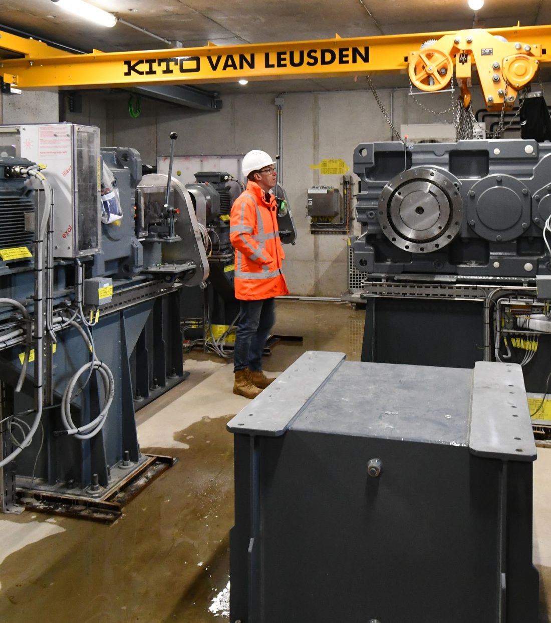 Ronald Smit in een technische ruimte met aandrijfsystemen, gekleed in oranje veiligheidsjas en helm